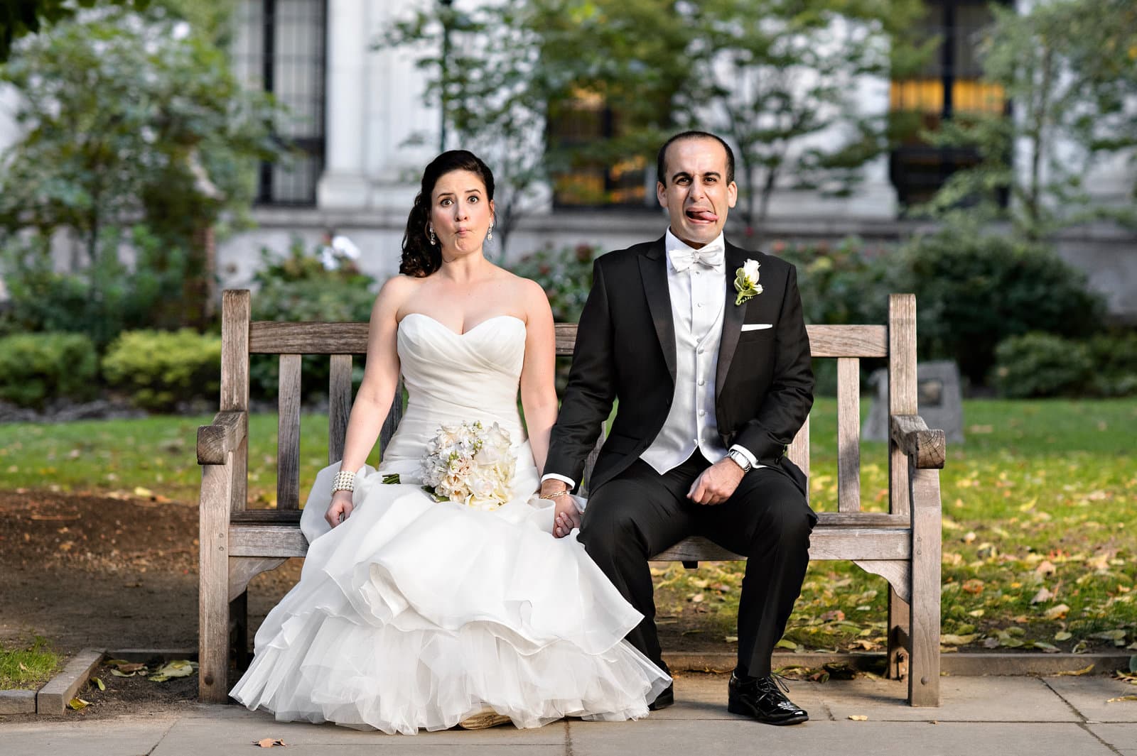 A bride and groom pulling silly faces on a park bench