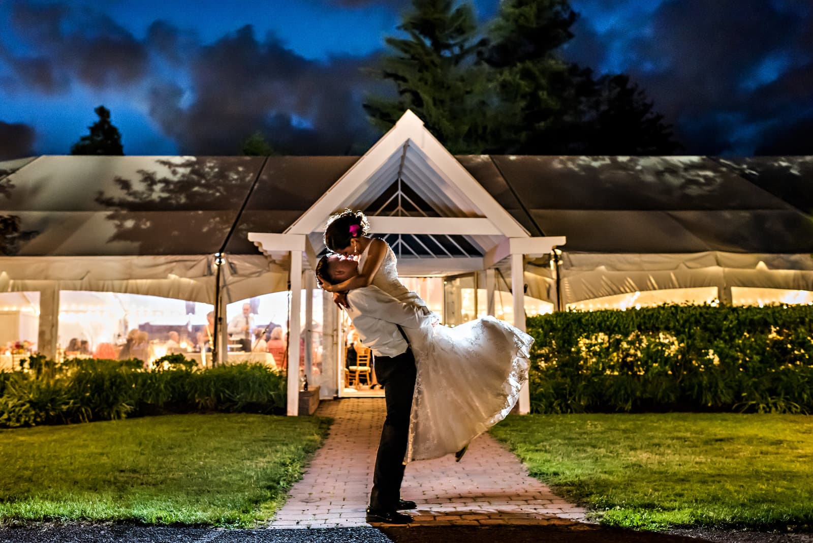 A couple dips into a kiss in front of a glowing reception tent at dusk