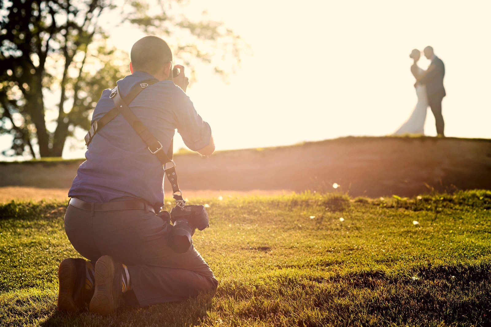 Russ Hickman low in the grass, photographing a couple at golden hour